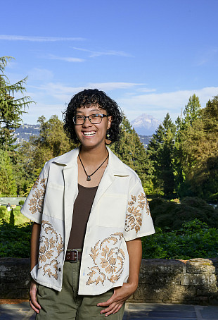 Meilin posing outside with Mount Hood in the background. They are wearing glasses and a white button-up shirt open over a black top.
