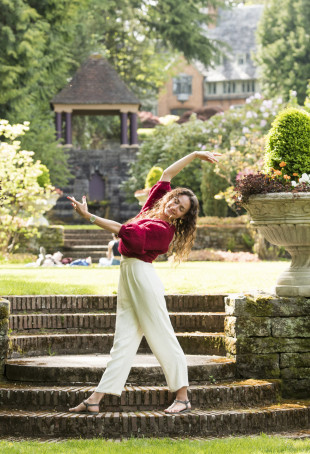 Katrina, in a red top and white pants, standing between two potted plants on brick stairs, eyes closed and arms outstretched into a plie ...